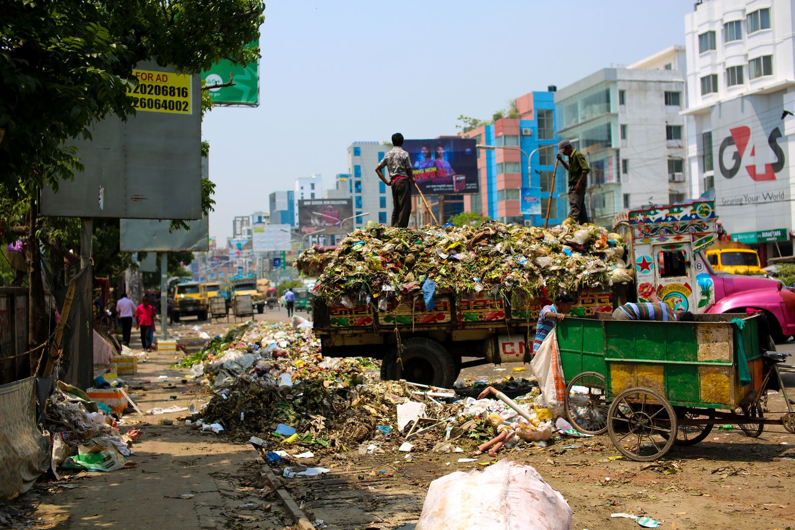 A city street with an overflowing waste truck, loose refuse spilling across the service lane, and waste pickers loading a handcart.